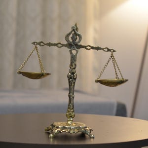 May include: A metallic gold-toned decorative scale of justice featuring an ornate central pillar with a decorative base, a horizontal crossbar, and two small, shallow bowls suspended by thin metal chains from each end of the crossbar.
