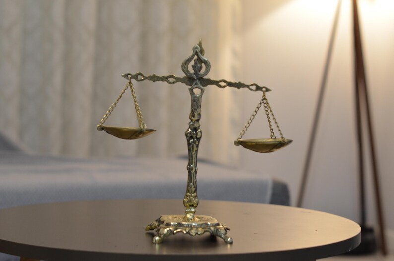 May include: A metallic gold-toned decorative scale of justice featuring an ornate central pillar with a decorative base, a horizontal crossbar, and two small, shallow bowls suspended by thin metal chains from each end of the crossbar.