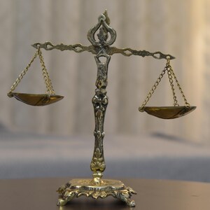 May include: A metallic scale of justice featuring an ornate central pillar with intricate scrollwork, a horizontal beam, and two suspended shallow bowls hanging from delicate gold-toned chains. The base is rectangular with decorative feet.
