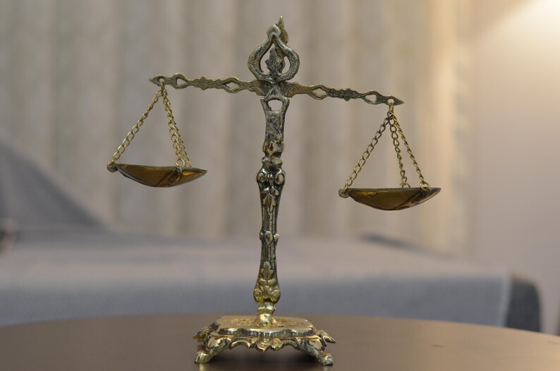 May include: A metallic scale of justice featuring an ornate central pillar with intricate scrollwork, a horizontal beam, and two suspended shallow bowls hanging from delicate gold-toned chains. The base is rectangular with decorative feet.
