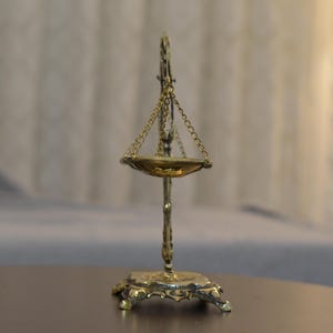 May include: A brass-colored metal scale featuring a central vertical stand with an ornate base, a single suspended shallow bowl held by three thin chains, and a decorative top finial, all displaying a metallic finish and intricate detailing.