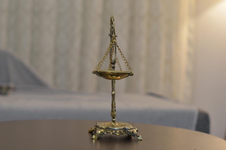 May include: A brass-colored metal scale featuring a central vertical stand with an ornate base, a single suspended shallow bowl held by three thin chains, and a decorative top finial, all displaying a metallic finish and intricate detailing.