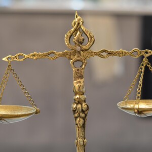 May include: A decorative brass balance scale featuring an ornate central pillar with intricate scrollwork and a top finial. Two shallow circular pans are suspended from the horizontal beam by thin metal chains on each side of the central support.