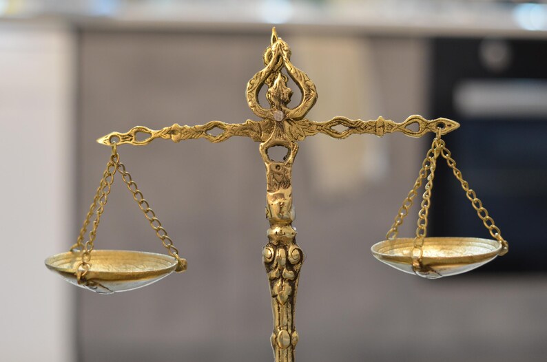 May include: A decorative brass balance scale featuring an ornate central pillar with intricate scrollwork and a top finial. Two shallow circular pans are suspended from the horizontal beam by thin metal chains on each side of the central support.