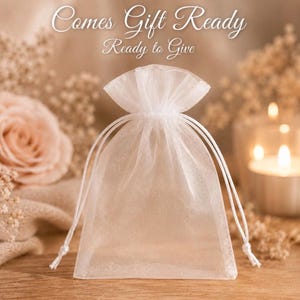 May include: A small, semi-transparent white organza drawstring pouch with a gathered top. The text "Comes Gift Ready" is printed in white script above the pouch. The bag is made of a sheer, lightweight fabric with matching white drawstrings.