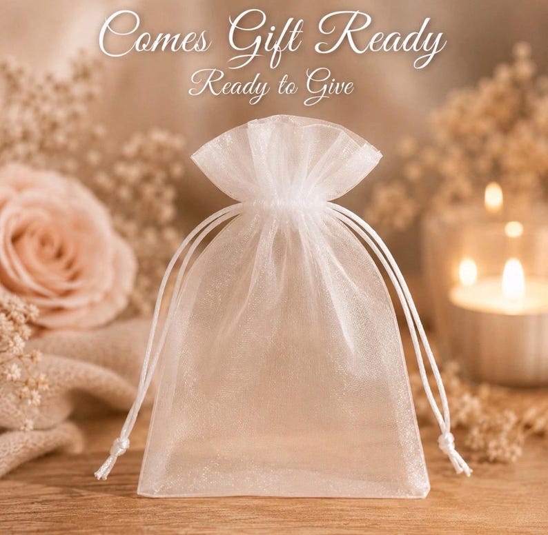 May include: A small, semi-transparent white organza drawstring pouch with a gathered top. The text "Comes Gift Ready" is printed in white script above the pouch. The bag is made of a sheer, lightweight fabric with matching white drawstrings.