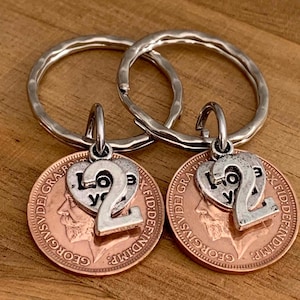 May include: Two silver-toned keychains featuring hammered metal rings. Each keychain holds a copper-colored coin charm with a profile portrait and the text GEORGIVS V DEI GRA BRITT OMN REX FID DEF IND IMP. A silver heart charm with the text LOVE YOU and a large number 2 are attached.