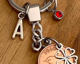 May include: A silver-toned metal keychain featuring a circular split ring, a letter A charm, a small red crystal charm, and a copper-colored 1946 farthing coin charm adorned with a silver-toned four-leaf clover cutout attached to the coin.