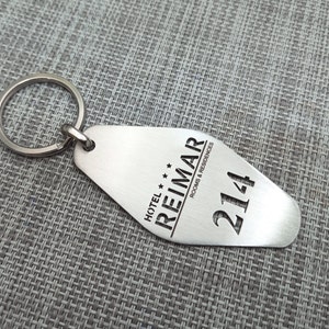 May include: A silver-colored hotel keychain with a key ring. The keychain has the text "HOTEL REIMAR ROOMS & RESIDENCES" and the number "214" cut out. The keychain is on a gray textured surface.