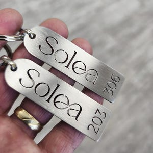 May include: Two brushed metal keychains with the name "Solea" cut out. One keychain has the number "306" and the other has "203" etched on it. The keychains are held by a person.