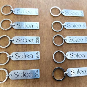 May include: A collection of silver rectangular keychains with the word "Solea" and a number engraved on each. The keychains have a circular ring for attaching keys. The background is a wooden surface.