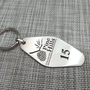 May include: A silver-colored metal keychain with a rounded end. The keychain has the words "Pine Hills Colorado" and the number "15" engraved on it, along with a pine cone design. A silver ring is attached.