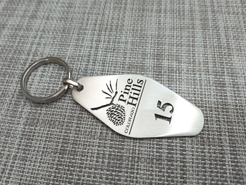 May include: A silver-colored metal keychain with a rounded end. The keychain has the words "Pine Hills Colorado" and the number "15" engraved on it, along with a pine cone design. A silver ring is attached.