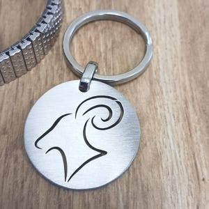 May include: Silver-toned keychain with a circular tag featuring a cut-out design of an Aries zodiac symbol. The keychain has a silver-toned ring.