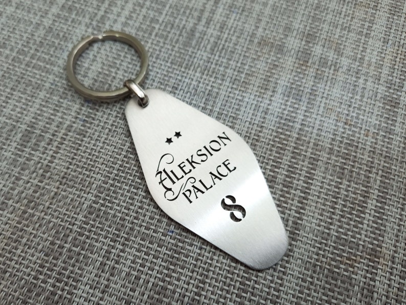 May include: A silver-colored metal keychain with a teardrop-shaped tag. The tag features the words "Aleksion Palace" in a decorative font, along with two stars and the number "8". A silver key ring is attached to the top.