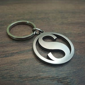 May include: A silver-colored keychain with a circular pendant featuring a stylized letter "S". The letter is cut out of the pendant, creating a negative space design. The keychain ring is also silver.