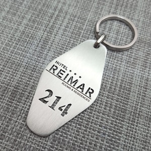 May include: A silver-colored hotel key tag with a key ring. The tag is shaped like a rounded rectangle and features the text "HOTEL REIMAR ROOMS & RESIDENCES" and the number "214".