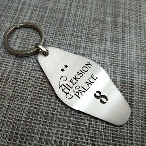 May include: A silver-colored metal keychain tag with the words "Aleksion Palace" and the number "8" etched on it. Two small stars are above the text. The keychain ring is attached to the top of the tag.