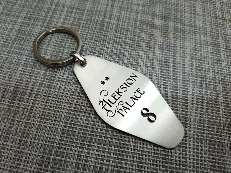 May include: A silver-colored metal keychain tag with the words "Aleksion Palace" and the number "8" etched on it. Two small stars are above the text. The keychain ring is attached to the top of the tag.
