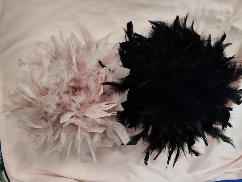 May include: Two feather embellishments, one in soft pink and the other in black. The pink one has a mix of pink and white feathers, while the black one is a dense cluster of dark feathers. Both are round in shape.