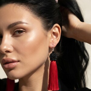 May include: A pair of long drop earrings featuring a vibrant red silk tassel suspended from a round, textured black beaded stud. The earrings are attached to silver-toned metal hooks and hang vertically, showcasing a contrast between the deep black beadwork and the bright red fringe.