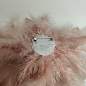 May include: A close-up of a light pink feather brooch. The brooch features a circular design with a cluster of soft, fluffy feathers. A silver pin is attached to the back for securing the brooch to clothing or accessories. The feathers are a delicate, pastel pink.