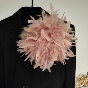 May include: A black blazer with a large, fluffy, light pink feather embellishment pinned to the lapel. The feathers are arranged in a dense, circular shape, creating a striking contrast against the dark fabric of the jacket. The blazer is hanging on a black hanger.