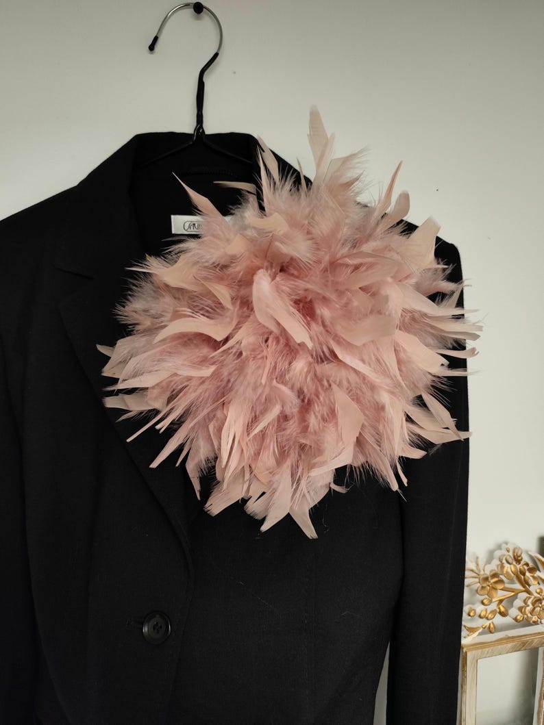 May include: A black blazer with a large, fluffy, light pink feather embellishment pinned to the lapel. The feathers are arranged in a dense, circular shape, creating a striking contrast against the dark fabric of the jacket. The blazer is hanging on a black hanger.