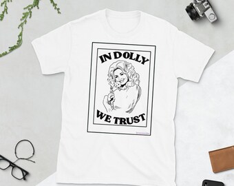 Könnte beinhalten: Ein weißes kurzärmeliges T-Shirt mit einem schwarz-weißen Grafikdruck einer Frau mit welligem Haar, die ein kleines Tier hält. Das rechteckige Design enthält den Text IN DOLLY WE TRUST in fetten schwarzen Großbuchstaben.
