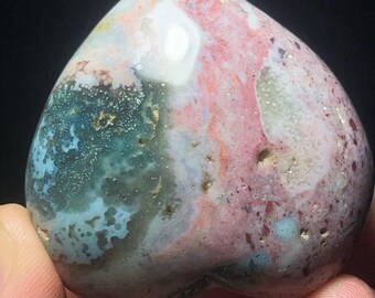 May include: A heart-shaped stone with a polished surface. The stone features a blend of colors, including pink, blue, green, and white, with a marbled appearance. The stone has a smooth, rounded shape and is held in a hand.