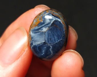 Könnte beinhalten: Ein ovaler Edelstein mit polierter Oberfläche. Der Stein hat eine tiefblaue Grundfarbe mit komplizierten weißen Mustern und orange-braunen Streifen. Der Edelstein wird in einer Hand vor dunklem Hintergrund gehalten.