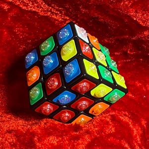 Può includere: Un cubo di Rubik nero con quadrati colorati, tra cui rosso, blu, verde, giallo e arancione. Ogni quadrato ha un centro trasparente a forma di gemma. Il cubo è posto su uno sfondo rosso vibrante.