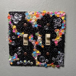 Op de afbeelding: Een decoratieve lichtschakelaar met twee schakelaars. De zwarte afdekking is versierd met een verscheidenheid aan kleurrijke, sprankelende edelstenen en kralen. De edelstenen zijn in tinten helder, roze, geel, oranje, blauw en groen.
