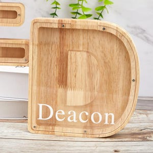 Peut inclure: Une tirelire en bois clair en forme de lettre D avec un panneau avant en acrylique transparent. Le nom Deacon est imprim&eacute; en lettres blanches majuscules et minuscules sur l'acrylique. De petites vis m&eacute;talliques fixent le panneau au cadre.