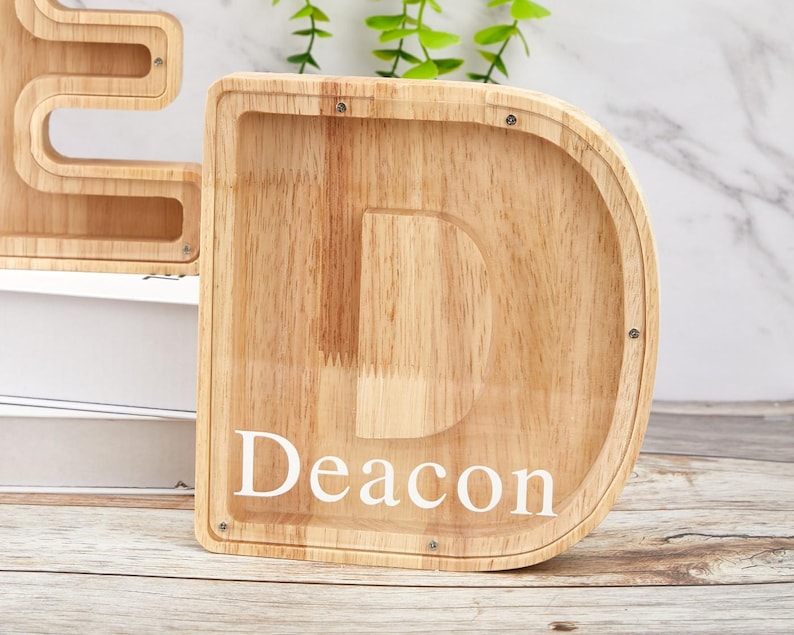 Peut inclure: Une tirelire en bois clair en forme de lettre D avec un panneau avant en acrylique transparent. Le nom Deacon est imprim&eacute; en lettres blanches majuscules et minuscules sur l'acrylique. De petites vis m&eacute;talliques fixent le panneau au cadre.
