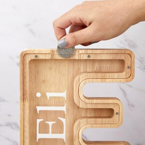 Peut inclure: Une tirelire en bois clair en forme de lettre majuscule E, dot&eacute;e d'une fa&ccedil;ade en acrylique transparent et du pr&eacute;nom Eli imprim&eacute; en blanc. Une pi&egrave;ce de monnaie argent&eacute;e est ins&eacute;r&eacute;e dans la fente sup&eacute;rieure du contenant en bois.