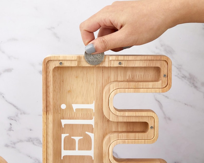 Peut inclure: Une tirelire en bois clair en forme de lettre majuscule E, dot&eacute;e d'une fa&ccedil;ade en acrylique transparent et du pr&eacute;nom Eli imprim&eacute; en blanc. Une pi&egrave;ce de monnaie argent&eacute;e est ins&eacute;r&eacute;e dans la fente sup&eacute;rieure du contenant en bois.