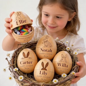 May include: Wooden Easter egg set with personalized names and bunny designs. The eggs are in a woven basket with flowers. One egg is open, revealing colorful candies. Each egg has "Happy Easter 2026" printed on it.