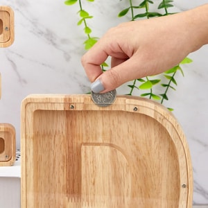 Peut inclure: Une tirelire en bois clair en forme de lettre D avec un panneau avant en acrylique transparent. Une pi&egrave;ce de monnaie argent&eacute;e est ins&eacute;r&eacute;e dans la fente &eacute;troite situ&eacute;e sur le dessus du cadre en bois, fix&eacute; par de petites vis m&eacute;talliques.