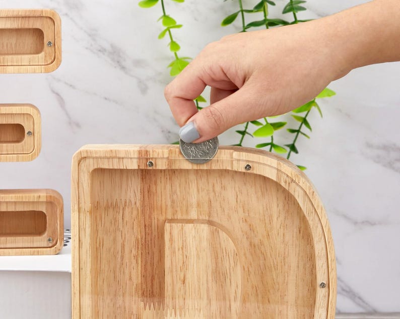 Peut inclure: Une tirelire en bois clair en forme de lettre D avec un panneau avant en acrylique transparent. Une pi&egrave;ce de monnaie argent&eacute;e est ins&eacute;r&eacute;e dans la fente &eacute;troite situ&eacute;e sur le dessus du cadre en bois, fix&eacute; par de petites vis m&eacute;talliques.