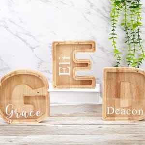 Peut inclure: Trois tirelires en bois en forme de lettres avec des fa&ccedil;ades en acrylique transparent. La lettre G porte le nom Grace, la lettre E le nom Eli et la lettre D le nom Deacon, tous imprim&eacute;s en blanc sur les surfaces transparentes.
