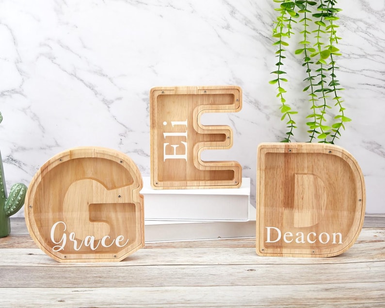 Peut inclure: Trois tirelires en bois en forme de lettres avec des fa&ccedil;ades en acrylique transparent. La lettre G porte le nom Grace, la lettre E le nom Eli et la lettre D le nom Deacon, tous imprim&eacute;s en blanc sur les surfaces transparentes.
