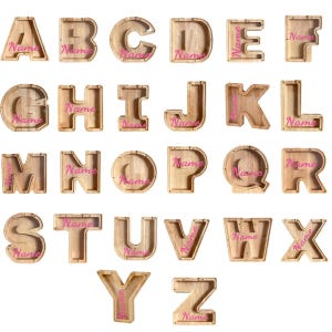 Peut inclure: Une collection de vingt-six tirelires en bois en forme de lettres, chacune dot&eacute;e d'une fa&ccedil;ade transparente et du mot Name imprim&eacute; en &eacute;criture rose. Les lettres de A &agrave; Z sont dispos&eacute;es en grille, r&eacute;v&eacute;lant le grain du bois clair naturel.