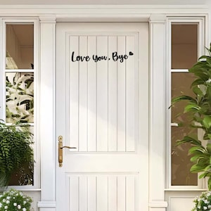 Pode incluir: Uma porta branca com painéis verticais e a inscrição preta "Love you, Bye". A porta tem uma maçaneta de latão e é emoldurada por molduras e janelas brancas. Plantas verdes são visíveis em ambos os lados da porta.