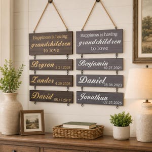 May include: Two vertical wall signs made of dark wood planks connected by metal rings and hung with rope. Each sign features the text "Happiness is having grandchildren to love" followed by three smaller planks displaying names and birth dates.