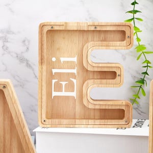 Peut inclure: Une tirelire en bois clair en forme de lettre majuscule E avec une fa&ccedil;ade en acrylique transparent. Le pr&eacute;nom Eli est imprim&eacute; en lettres capitales blanches sur la surface transparente. Le cadre en bois est fix&eacute; par de petites vis m&eacute;talliques.