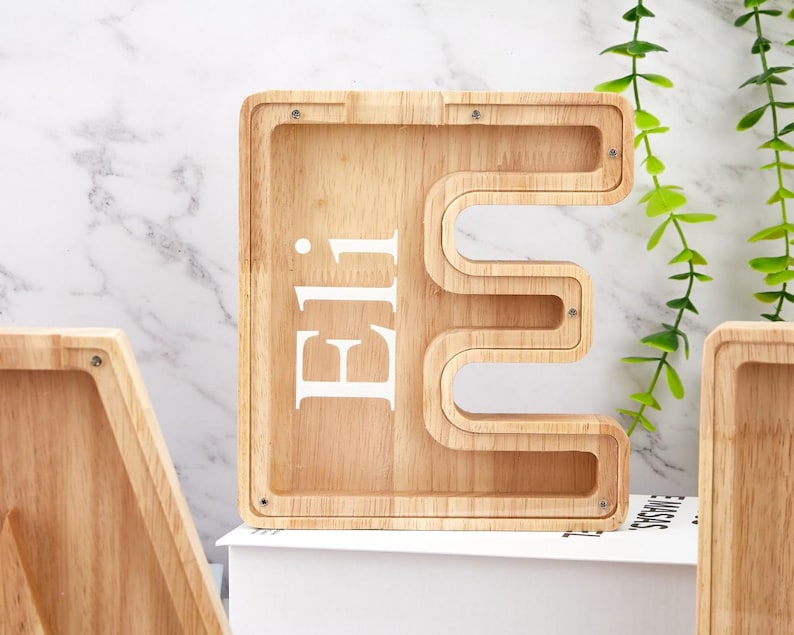 Peut inclure: Une tirelire en bois clair en forme de lettre majuscule E avec une fa&ccedil;ade en acrylique transparent. Le pr&eacute;nom Eli est imprim&eacute; en lettres capitales blanches sur la surface transparente. Le cadre en bois est fix&eacute; par de petites vis m&eacute;talliques.