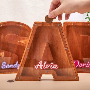 May include: Wooden letter-shaped piggy banks with clear acrylic fronts. The letter "A" is in focus, with the name "Alvin" in red script. A hand is dropping a coin into the slot. Other letters are visible, with names "Sandy" and "Doris".
