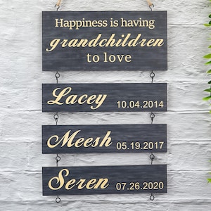 Puede incluir: Cuatro letreros de madera cuelgan de una cuerda. El letrero superior dice "Happiness is having grandchildren to love". Los tres letreros de abajo enumeran nombres y fechas: "Lacey 10.04.2014", "Meesh 05.19.2017" y "Seren 07.26.2020".