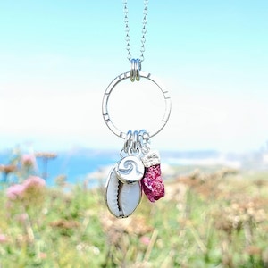 May include: A silver necklace with a large circular pendant and three charms: a white seashell, a pink crystal, and a silver spiral. The necklace hangs against a blurred backdrop of the sea and greenery.